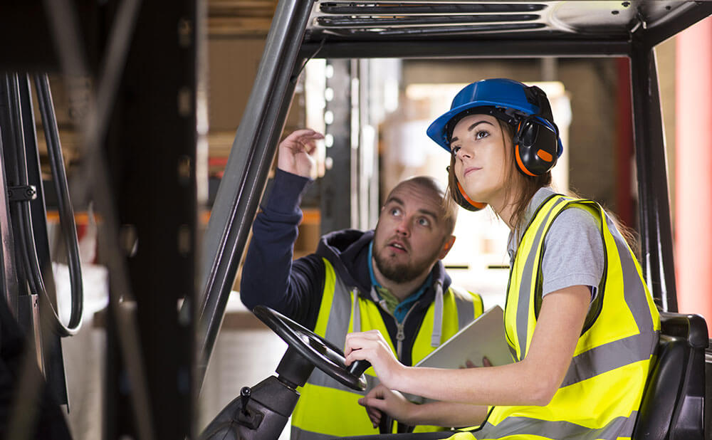 Sécurité au travail Une femme conduit un charriot élévateur et un homme à côté une explique quelque chose. Ils portent tous deux un gilet jaune. La femme porte en plus un casque et casque anti bruit sur ses oreilles.